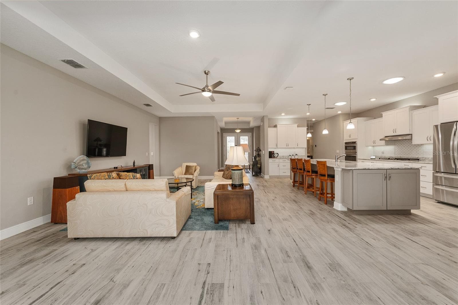 A stunning view of the spacious great room and beautifully appointed kitchen, showcasing an elegant tray ceiling, ceramic plank flooring, recessed lighting, and two kitchen solar tubes that bring additional natural light into the space. - 8290 SW 92ND CIR, OCALA