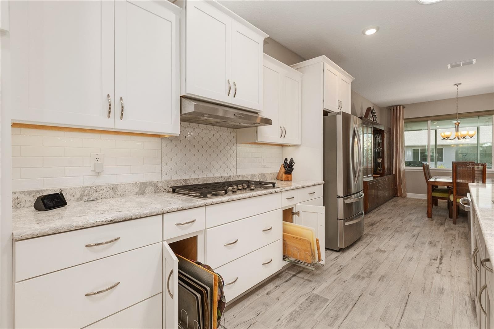 Thoughtfully designed kitchen featuring convenient pull-out storage organizers for cutting boards and baking sheets, Cambria quartz countertops, decorative tile backsplash, over- and under-cabinet lighting, stainless steel appliances, and an open flow into the dining area. - 8290 SW 92ND CIR, OCALA