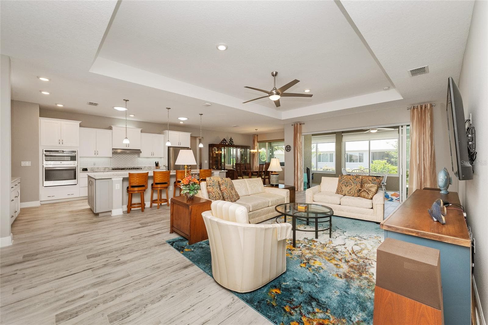 Spacious open-concept great room featuring a tray ceiling, recessed lighting, ceramic plank flooring, and stacking sliding glass doors with a zero-threshold entry to the enclosed lanai, creating a seamless transition between indoor and outdoor living spaces. - 8290 SW 92ND CIR, OCALA