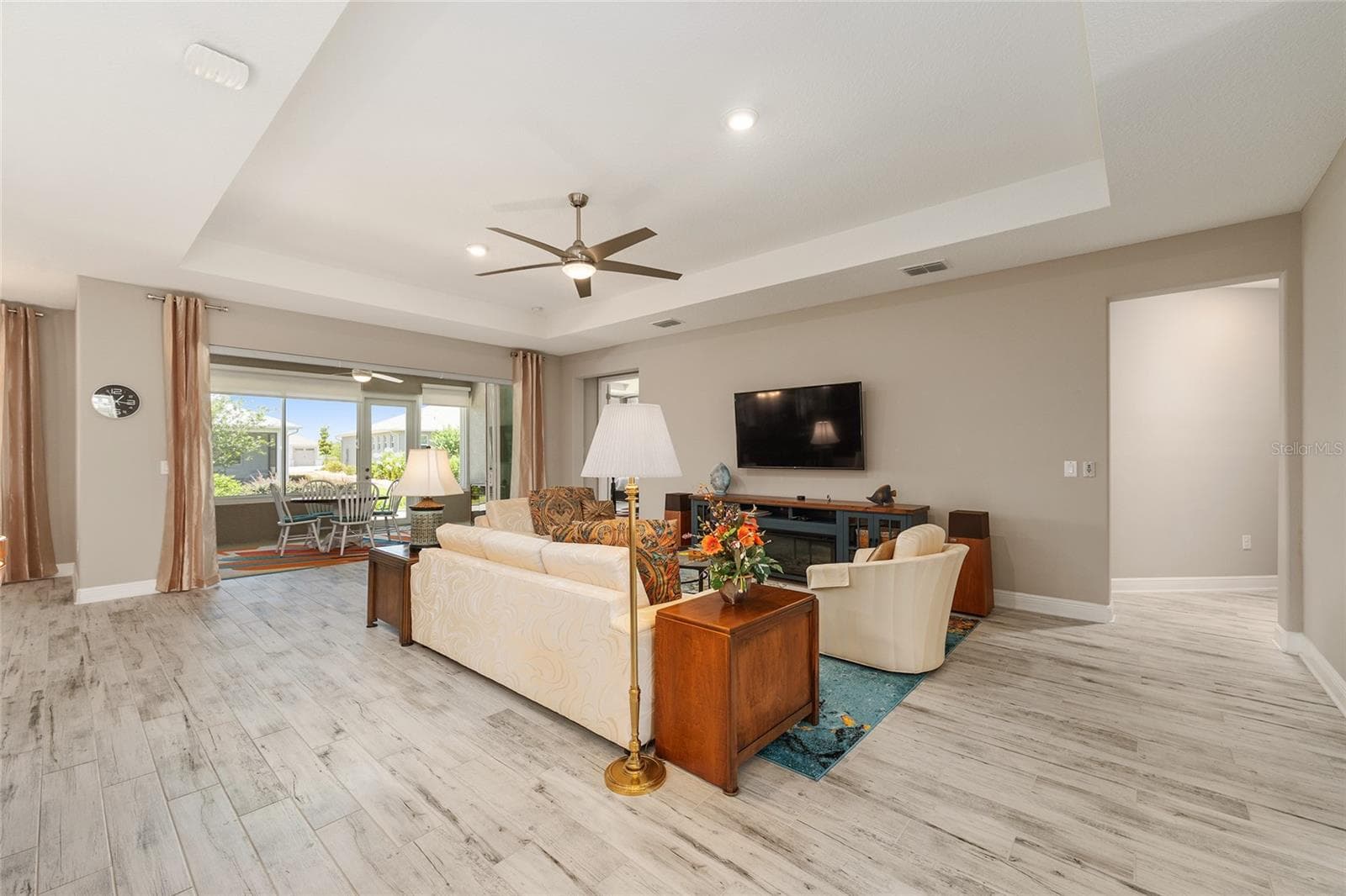 Expansive great room featuring a tray ceiling, recessed lighting, ceiling fan with integrated lighting, ceramic plank flooring, and stacking sliding glass doors with a zero-threshold transition to the enclosed lanai. An inviting space for relaxing and entertaining, with the wall-mounted TV included in the sale. - 8290 SW 92ND CIR, OCALA