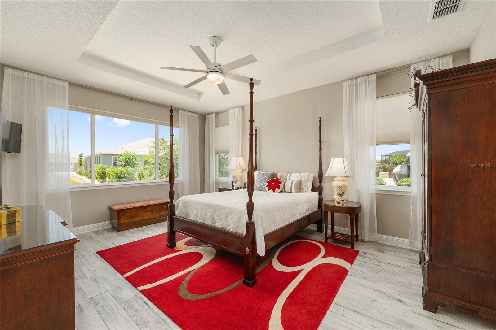 Spacious first primary suite, currently used as the owner's suite, featuring an elegant tray ceiling, ceiling fan with integrated lighting, ceramic plank flooring, neutral finishes, and multiple windows that fill the room with natural light. Wall-mounted TV included in the sale. - 8290 SW 92ND CIR, OCALA
