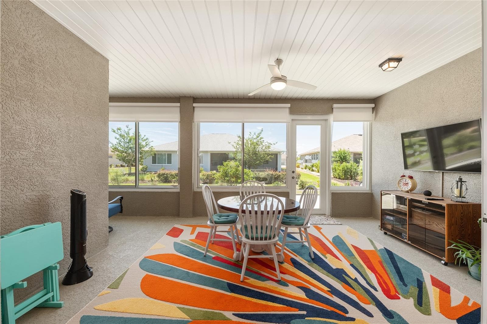 Inviting enclosed lanai featuring custom solar shades, a ceiling fan with integrated lighting, and expansive windows that fill the space with natural light. A perfect setting for relaxing or entertaining, with the wall-mounted TV included in the sale. - 8290 SW 92ND CIR, OCALA