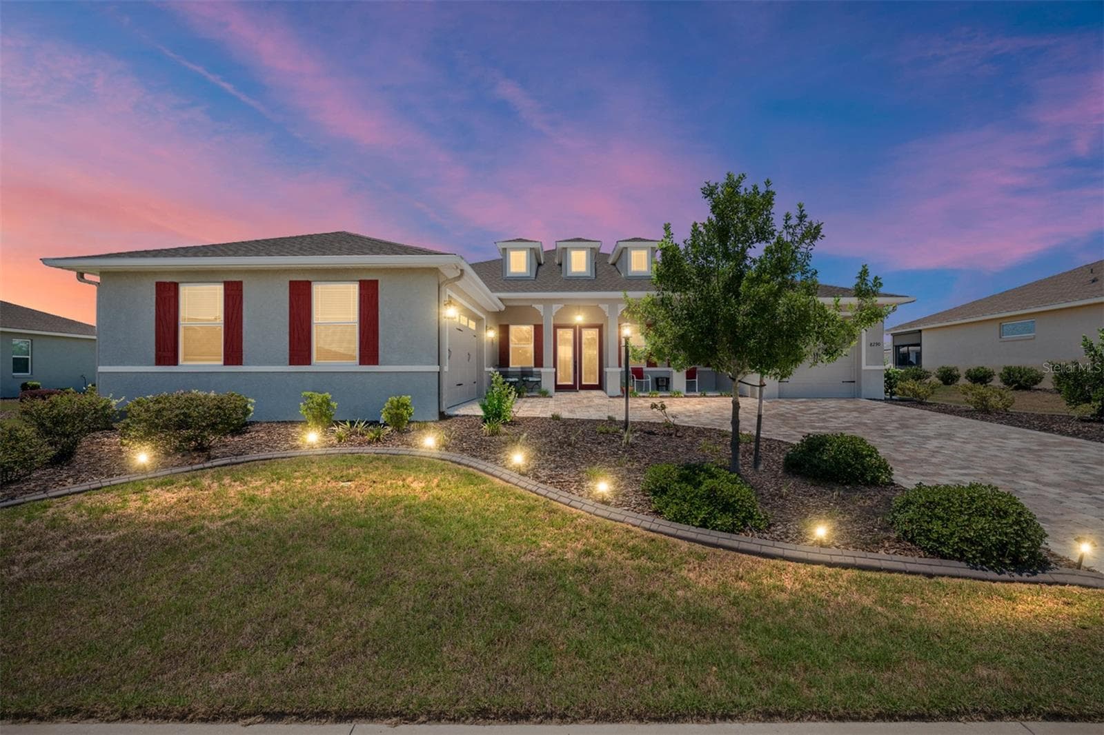 Stunning twilight view highlighting the home's exceptional curb appeal, beautifully illuminated landscaping, expansive paver driveway, and welcoming covered front porch. The elegant leaded-glass double entry doors and warm exterior lighting create an inviting first impression. - 8290 SW 92ND CIR, OCALA