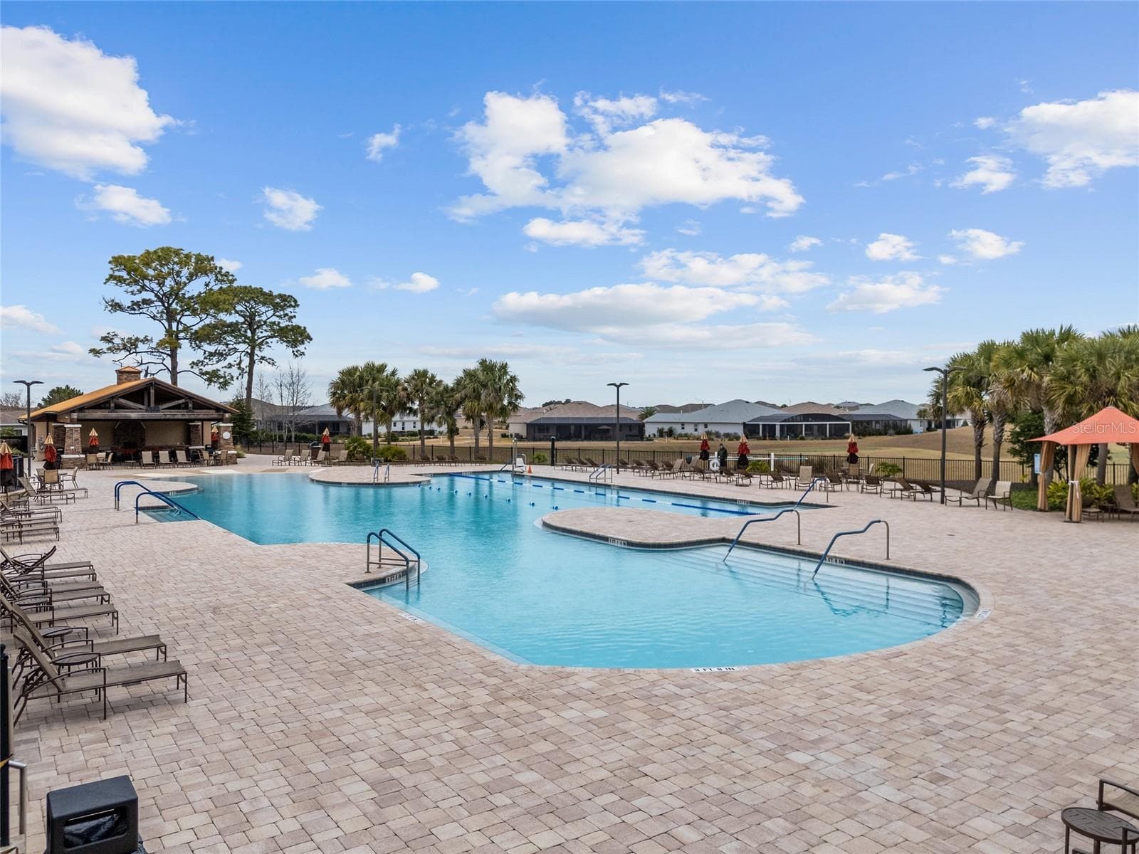 Another beautiful view of the Candler Hills resort-style heated pool, showcasing the expansive sun deck, abundant lounge seating, tropical landscaping, and covered pavilion—an inviting setting for soaking up the Florida sunshine and socializing with neighbors. - 8290 SW 92ND CIR, OCALA