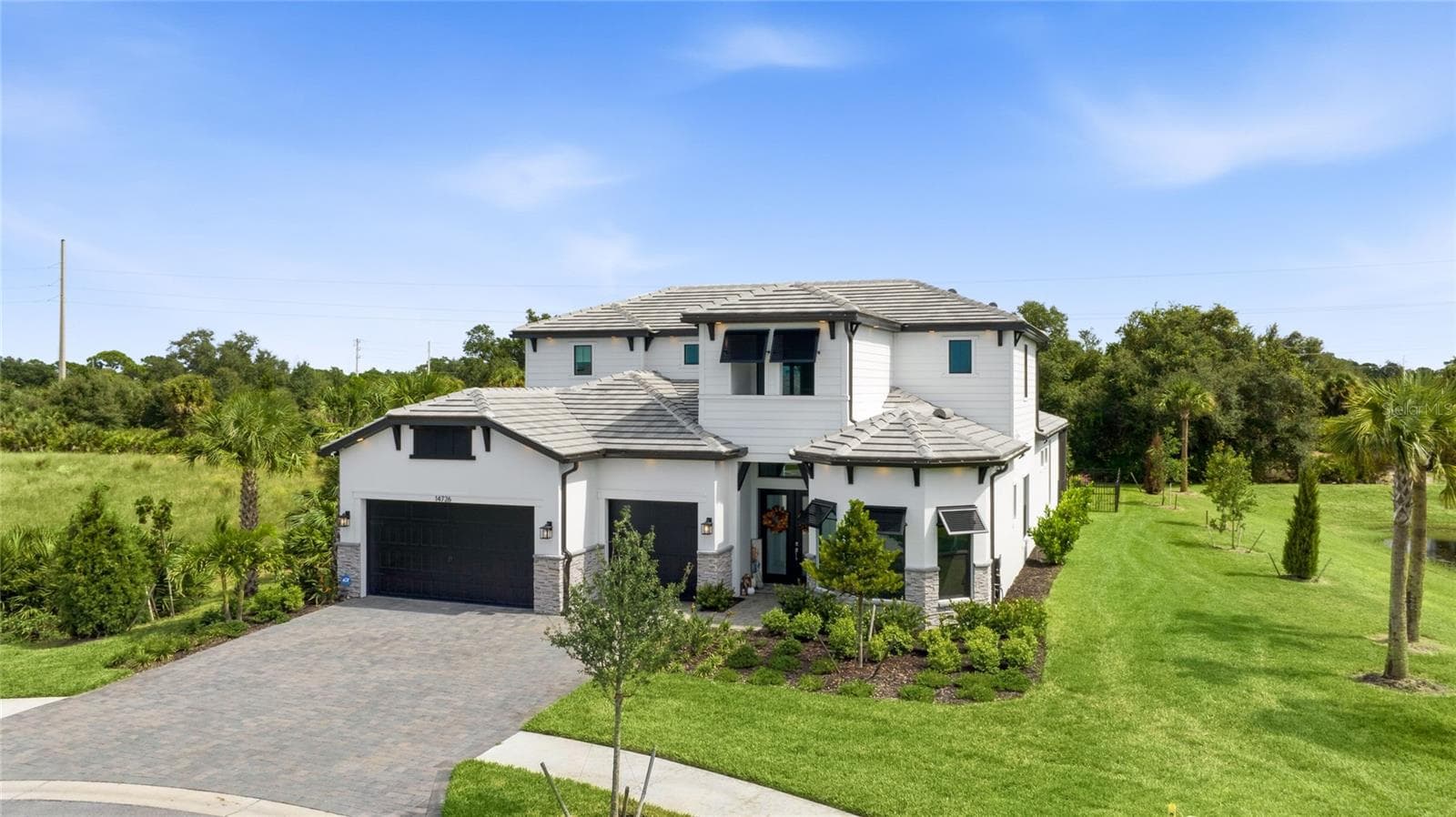 Elegant two-story residence featuring a striking modern coastal exterior, lush tropical landscaping, and an inviting paver driveway leading to a spacious three-car garage. - 14726 EAGLE BRANCH DR, NOKOMIS