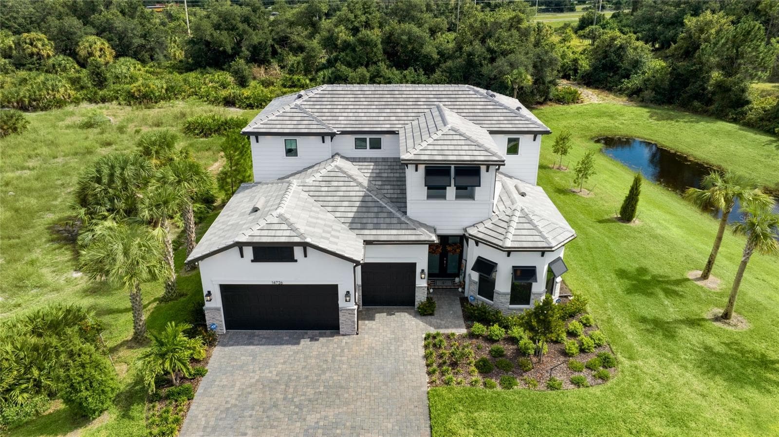 Elegant two-story residence featuring a striking modern coastal exterior, lush tropical landscaping, and an inviting paver driveway leading to a spacious three-car garage. - 14726 EAGLE BRANCH DR, NOKOMIS