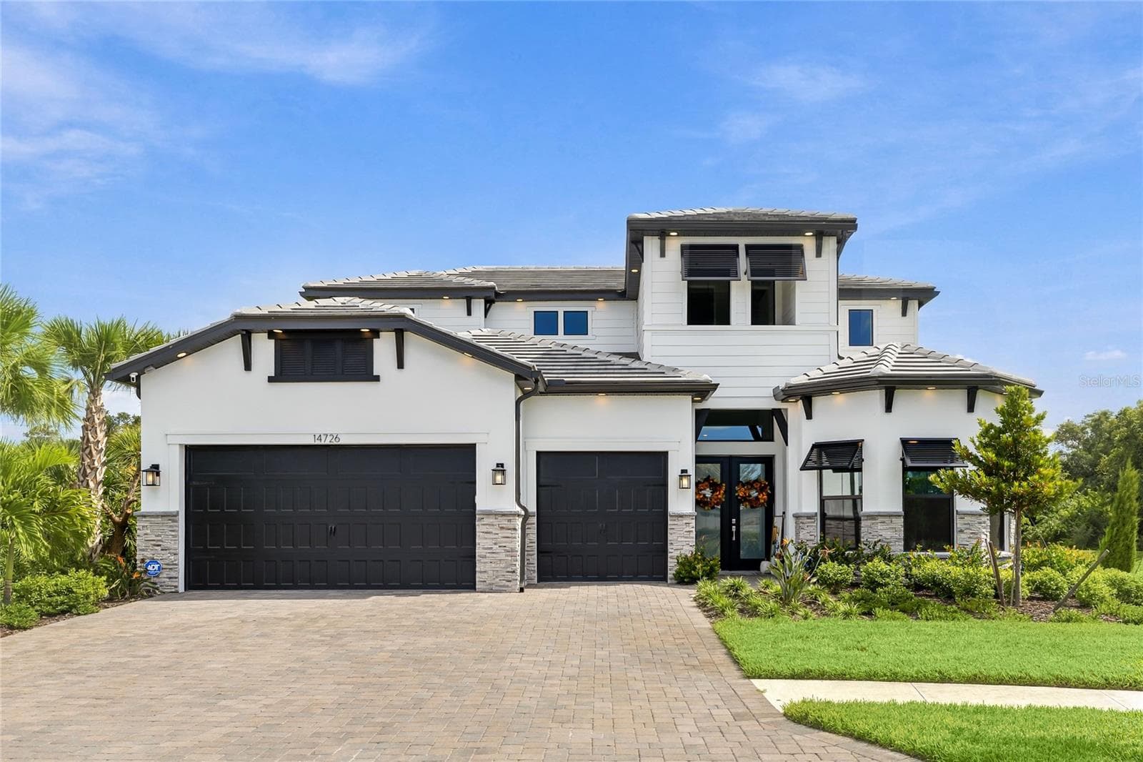 Elegant two-story residence featuring a striking modern coastal exterior, lush tropical landscaping, and an inviting paver driveway leading to a spacious three-car garage. - 14726 EAGLE BRANCH DR, NOKOMIS