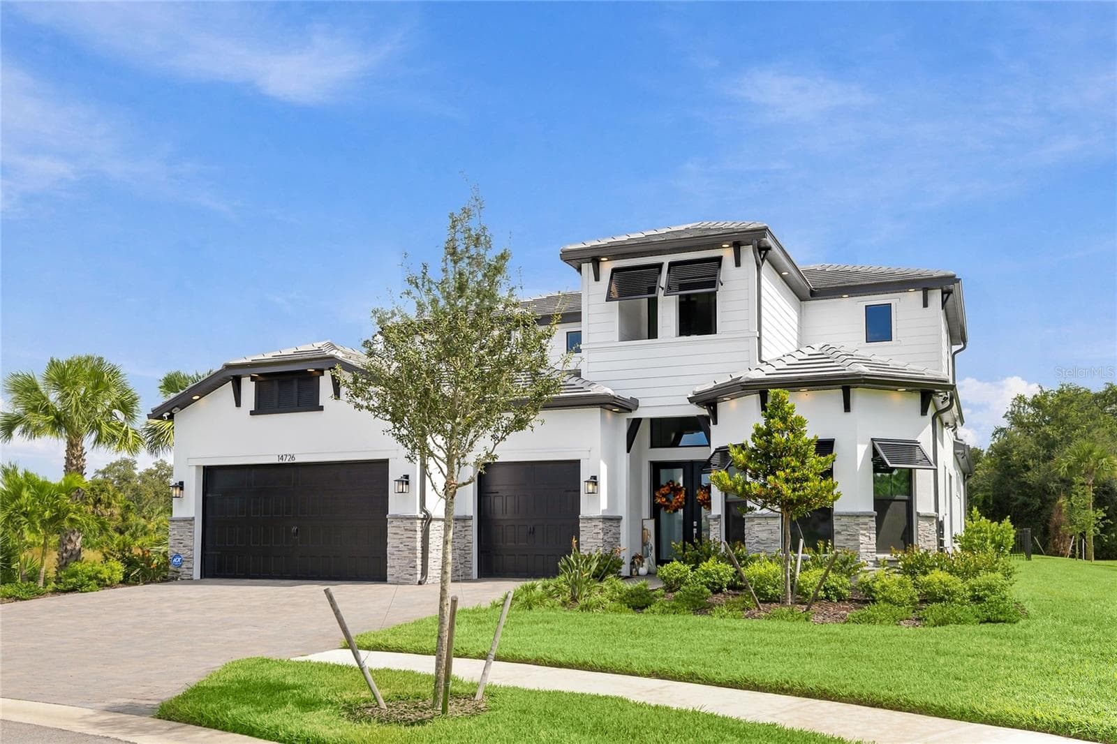 Elegant two-story residence featuring a striking modern coastal exterior, lush tropical landscaping, and an inviting paver driveway leading to a spacious three-car garage. - 14726 EAGLE BRANCH DR, NOKOMIS
