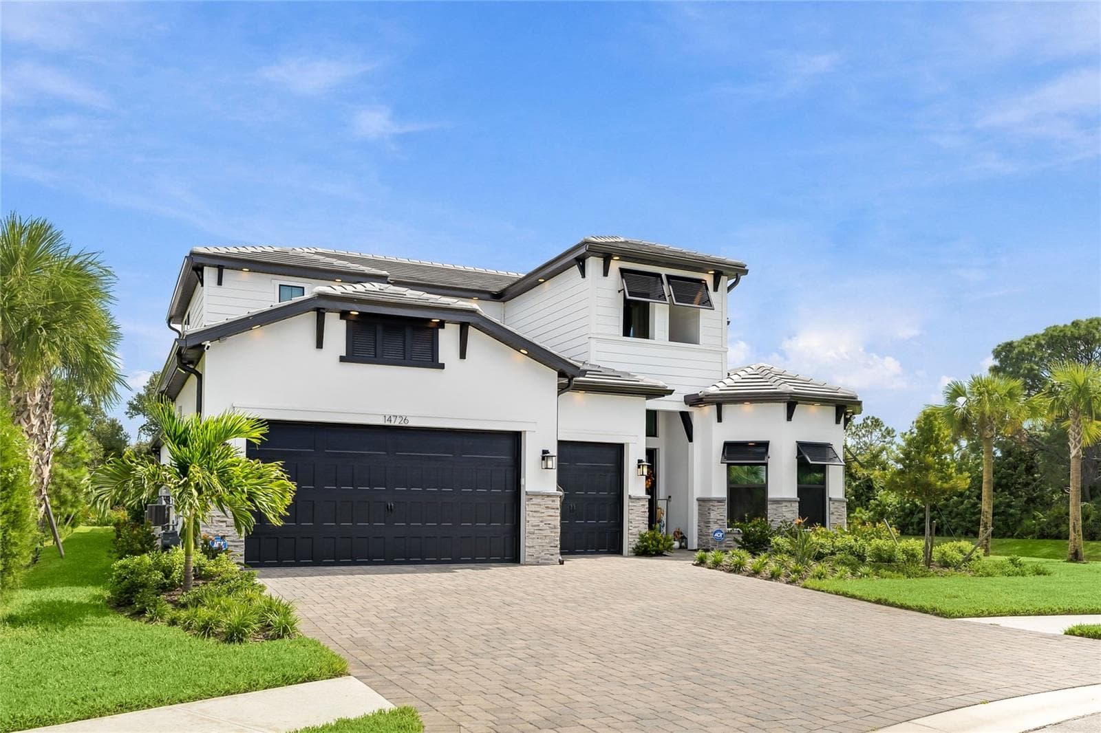 Elegant two-story residence featuring a striking modern coastal exterior, lush tropical landscaping, and an inviting paver driveway leading to a spacious three-car garage. - 14726 EAGLE BRANCH DR, NOKOMIS