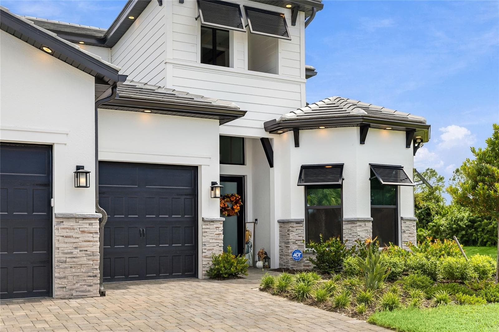 Elegant two-story residence featuring a striking modern coastal exterior, lush tropical landscaping, and an inviting paver driveway leading to a spacious three-car garage. - 14726 EAGLE BRANCH DR, NOKOMIS
