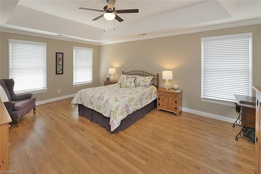 Inviting primary en suite featuring elegant tray ceiling, ceiling fan, gleaming hardwood floors, and an abundance of natural light. - 1101 Clyde Edgerton Drive, Kernersville NC 27284, Kernersville