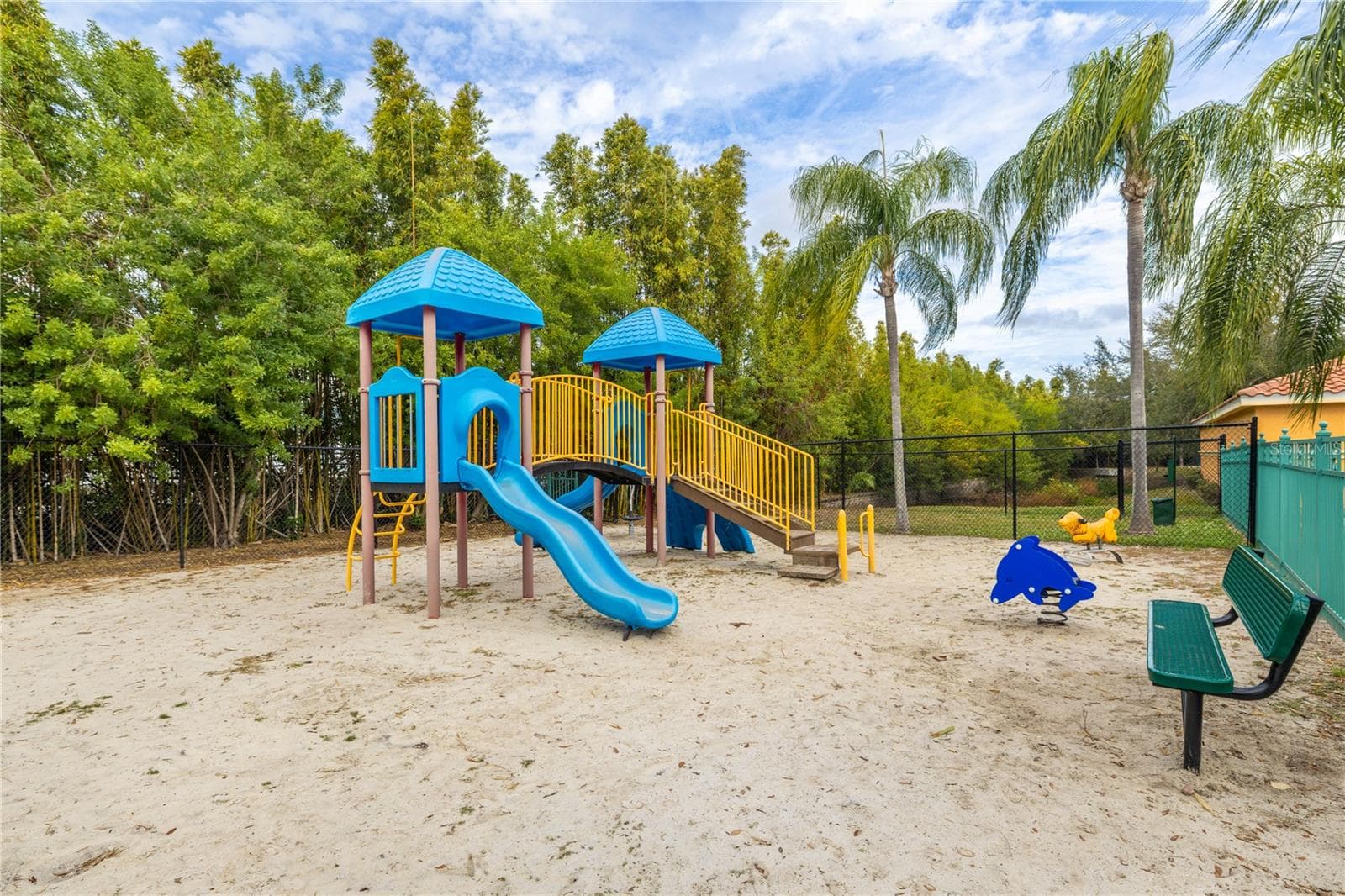 Community playground with a shaded play structure, slides, and seating, set in a sandy play area surrounded by tropical landscaping—perfect for family-friendly fun. - 2738 VIA TIVOLI #220B, CLEARWATER