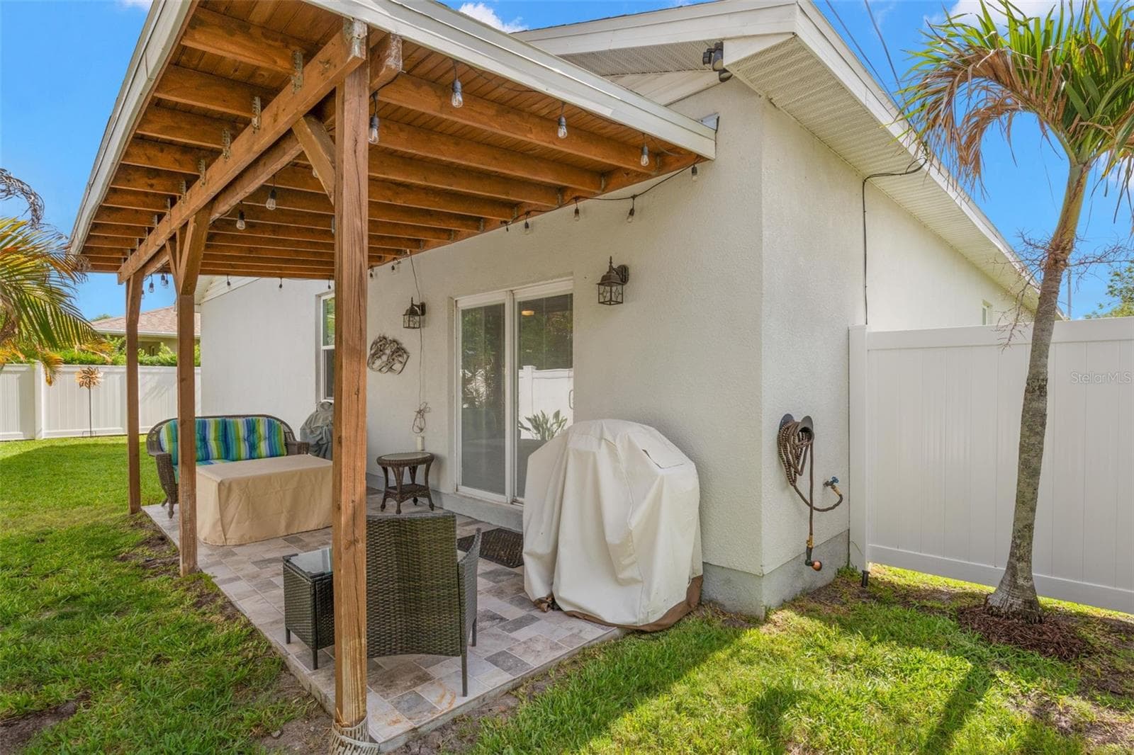 The covered rear patio extends the living space outdoors with a warm wood ceiling structure, decorative string lighting, ceramic tile flooring, and room for both dining and lounging. Sliding glass doors provide easy access to the interior, while vinyl fencing creates privacy throughout the backyard. - 1720 VERDE DR, CLEARWATER
