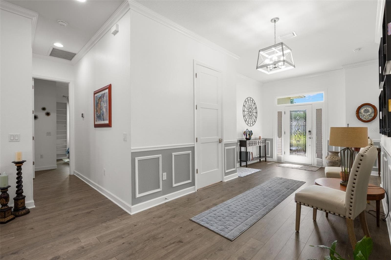 The oversized foyer is framed by extensive crown molding, oversized base trim, wainscoting, chair rail detailing, recessed lighting, and wide-plank luxury vinyl flooring. A statement chandelier, elegant leaded glass entry door with sidelights and transom window, and oversized paneled interior doors contribute to the home’s polished architectural character. - 1720 VERDE DR, CLEARWATER