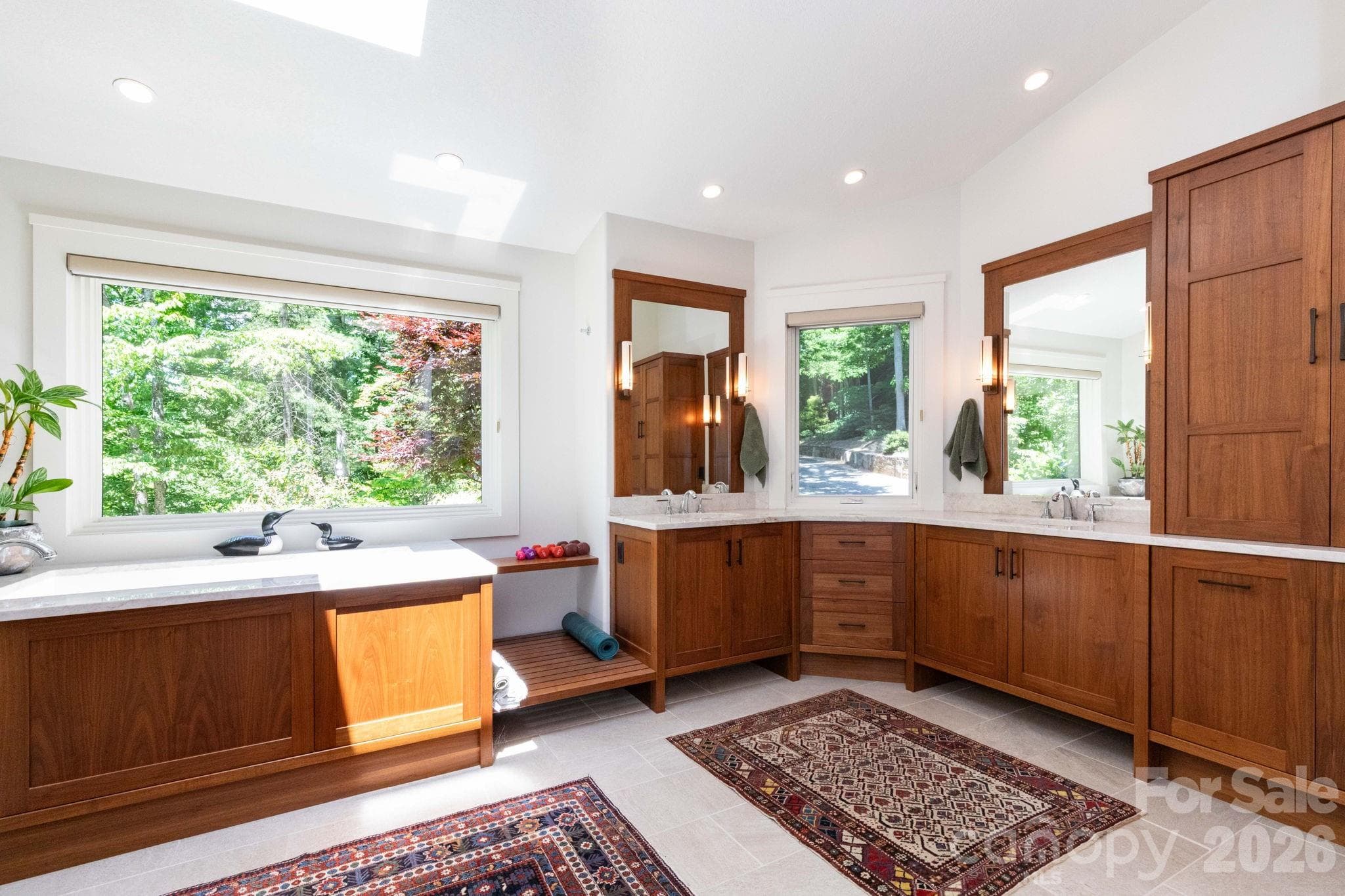 Spa-inspired primary bath featuring custom cabinetry, dual vanities, soaking tub, skylight, and oversized windows overlooking the lush surrounding landscape. - 29 Gardner Drive, Asheville