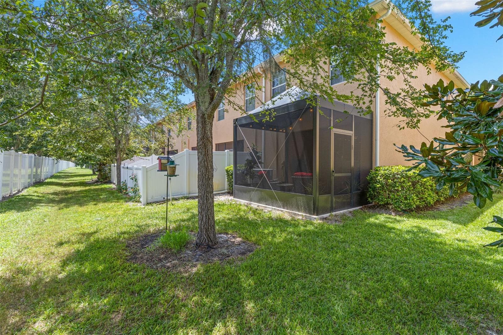 From the rear, the 2026 screened lanai sits beneath a canopy of mature trees, with a grassy common area adding space and greenery beyond. - 9524 TRUMPET VINE LOOP, NEW PORT RICHEY