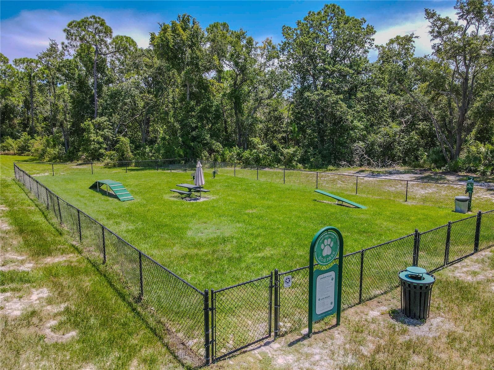 The community dog park provides a large fenced grassy area with agility equipment, bench seating and a shaded picnic table, all set against a wooded backdrop. - 3029 JACOB CROSSING LN, HOLIDAY