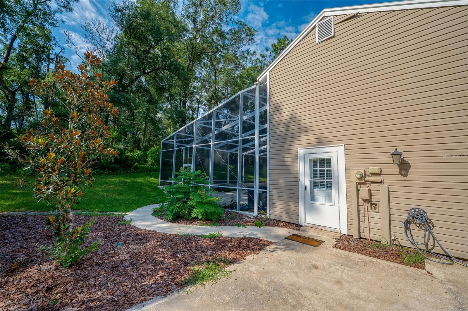Rear exterior view of the home featuring tan vinyl siding, a paved walkway, landscaped mulch beds, and an attached screened-in enclosure. - 2081 PENNSYLVANIA DR, DELAND