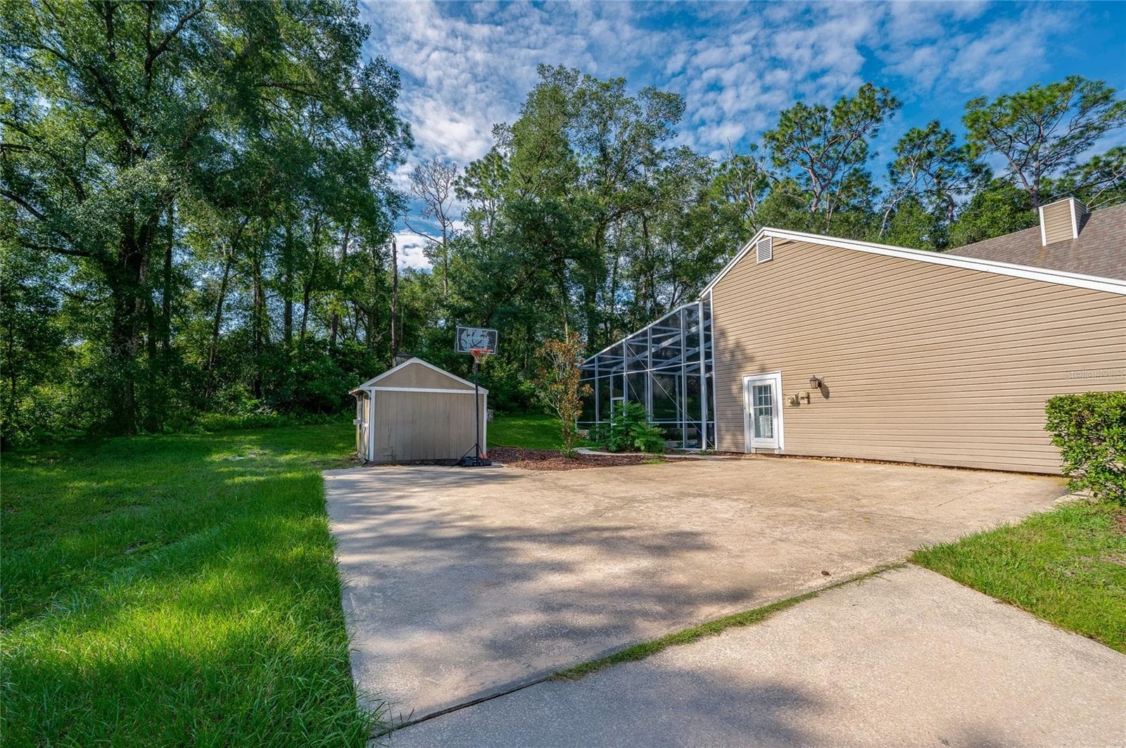 Open backyard space featuring a large concrete driveway area, an outdoor basketball hoop, a storage shed, and surrounding lush greenery. - 2081 PENNSYLVANIA DR, DELAND