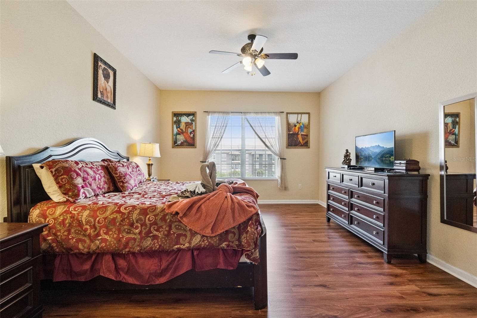Primary Suite Overview:Expansive primary bedroom featuring rich wood-look flooring, dark wood furnishings, ceiling fan, flat-screen TV console, and a large window providing abundant natural light. - 4840 CAYVIEW AVE #405, ORLANDO