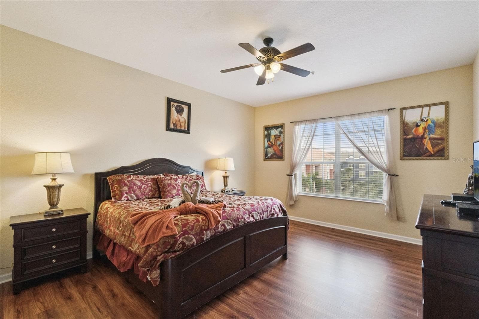 Primary Suite Layout:Spacious primary retreat displaying warm neutral tones, wood-look plank flooring, matching dark wood headboard and nightstands, ceiling fan, and a bright window. - 4840 CAYVIEW AVE #405, ORLANDO