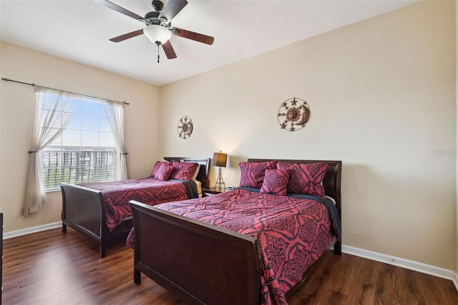 Guest Bedroom Window View:Light-filled bedroom space featuring two dark wood bed frames, central nightstand with lamp, ceiling fan, warm neutral walls, and wood-look plank flooring. - 4840 CAYVIEW AVE #405, ORLANDO
