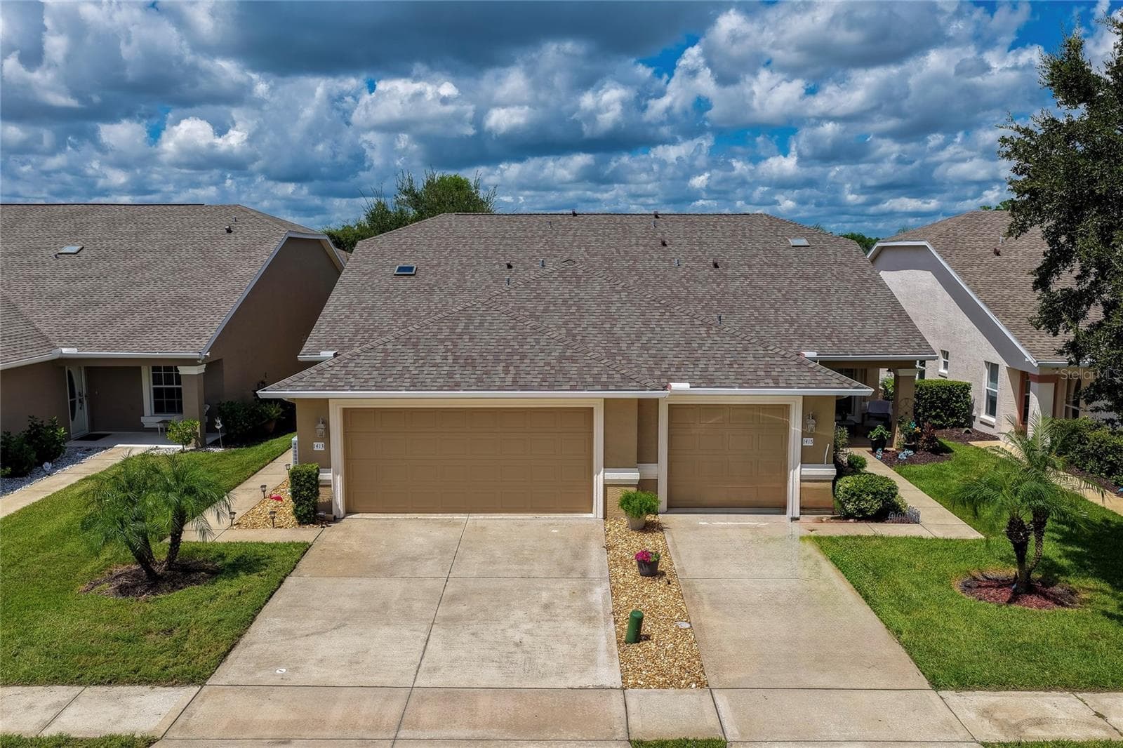 Front-facing view of the home featuring neutral stucco siding, an attached garage, and an expansive paved driveway surrounded by tropical green landscaping. - 1413 COCONUT PALM CIR, PORT ORANGE