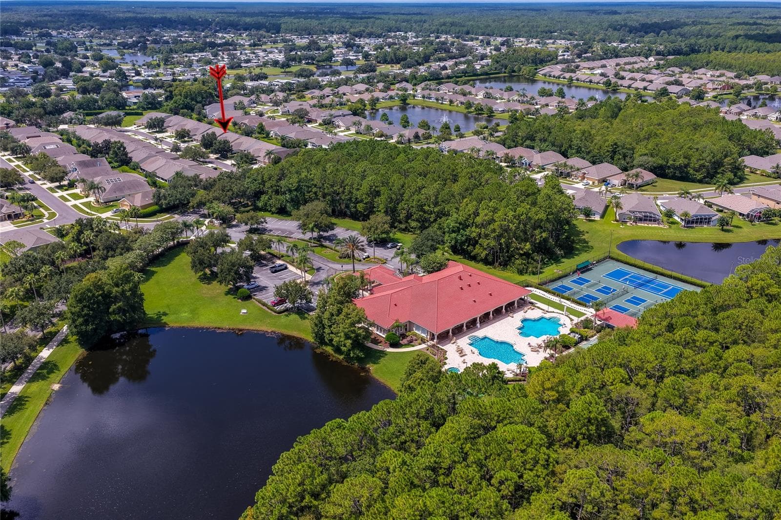 Elevated wide-angle aerial perspective highlighting the community clubhouse, sports courts, and adjacent lake, with an arrow pointing toward the neighborhood homes nestled behind green tree lines. - 1413 COCONUT PALM CIR, PORT ORANGE