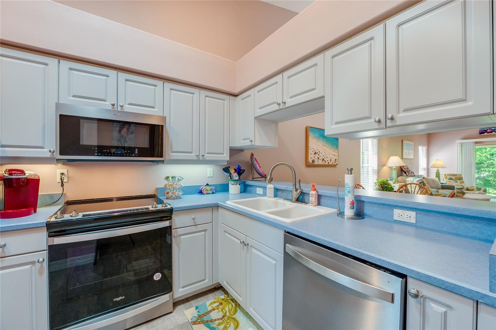 Efficient kitchen layout highlighting a built-in dishwasher, electric range, microwave, and double-basin sink. A pass-through window maintains a seamless connection to the adjacent living area. - 1413 COCONUT PALM CIR, PORT ORANGE