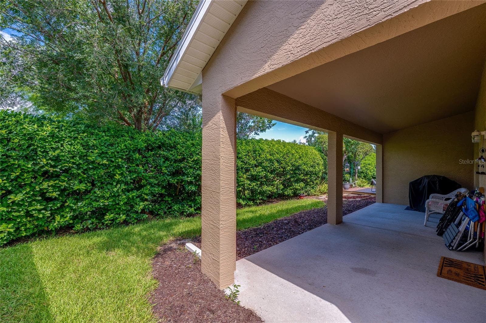 Covered concrete back patio offering protection from the elements alongside a manicured green lawn. Tall, dense privacy hedges frame the perimeter to create a quiet outdoor sanctuary. - 1413 COCONUT PALM CIR, PORT ORANGE