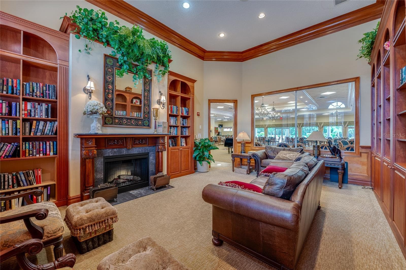 Warmly styled library corner highlighted by a classic wood-mantel fireplace with a stone surround, vibrant wall greenery, and custom built-in shelving. A large interior window cut-out looks directly through into the main event hall. - 1413 COCONUT PALM CIR, PORT ORANGE