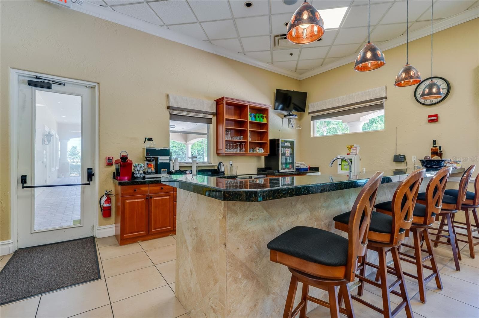 Clean service bar area featuring dark granite counter surfaces, warm wood cabinetry, neutral tile flooring, and industrial-style hanging pendant lights. An exterior door opens directly to a paved covered walkway outside. - 1413 COCONUT PALM CIR, PORT ORANGE