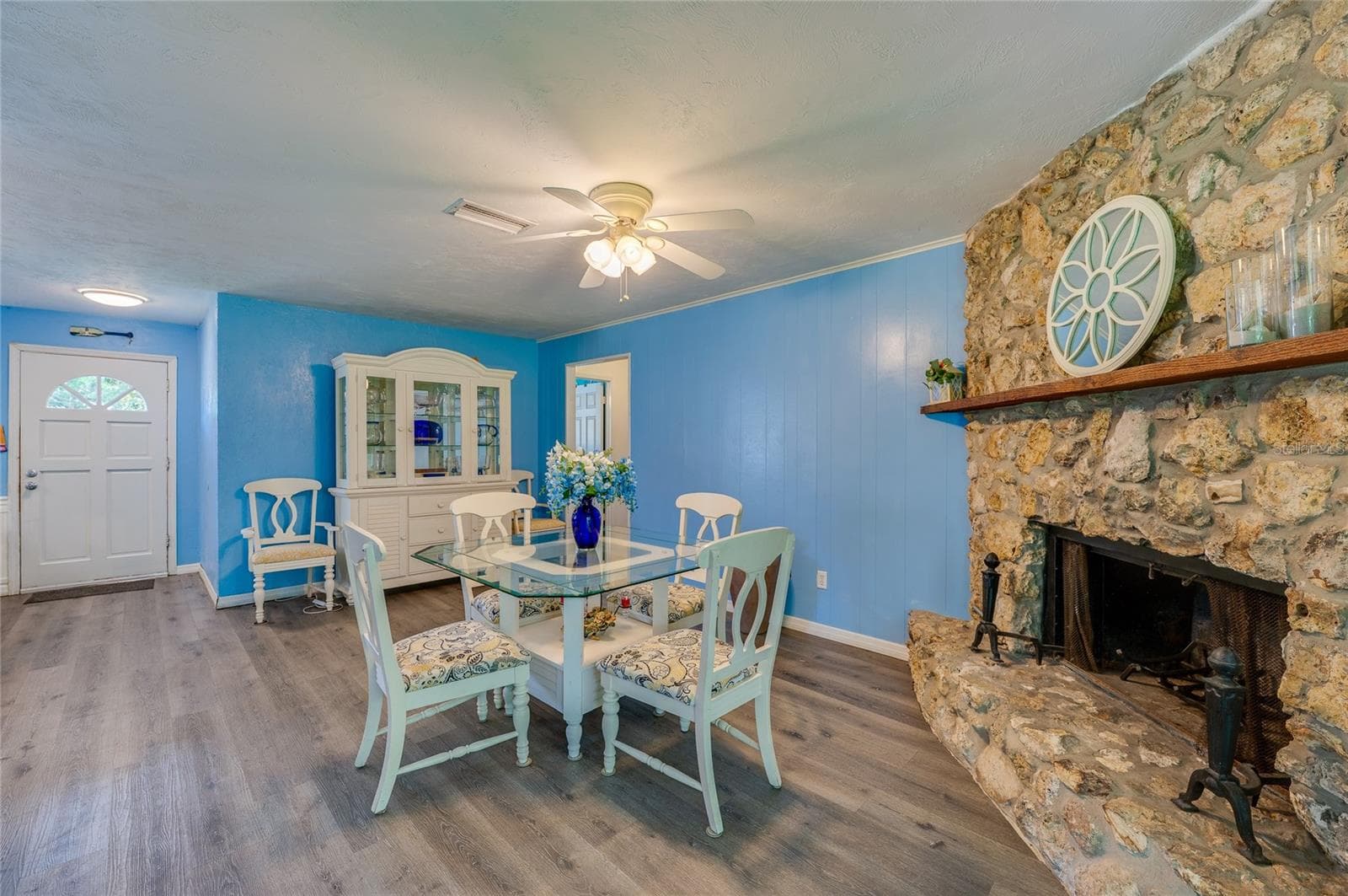 Open-concept living room anchored by a rustic stone fireplace, featuring light blue walls, LVP flooring, and a lighted ceiling fan. - 146 WESTWOOD DR, DAYTONA BEACH