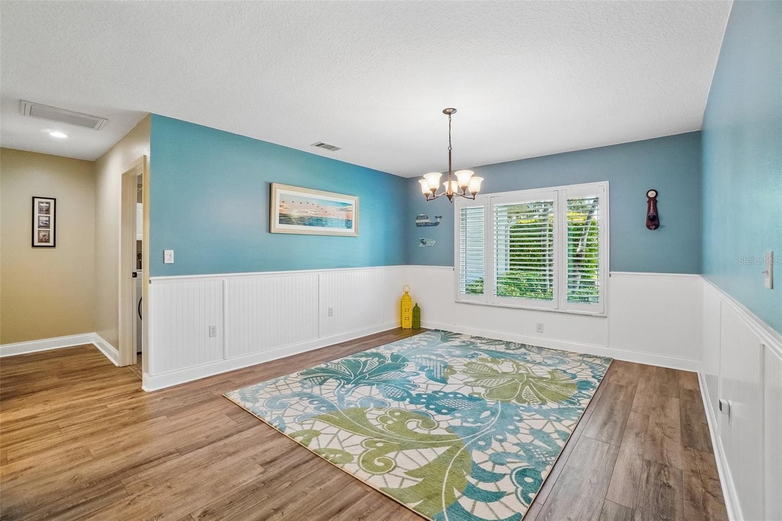 Dining Room – This flexible dining space features wood-look flooring, white beadboard wainscoting, a decorative chandelier, and a large plantation-shuttered window overlooking the greenery outside. - 971 CORTLAND WAY, PALM HARBOR