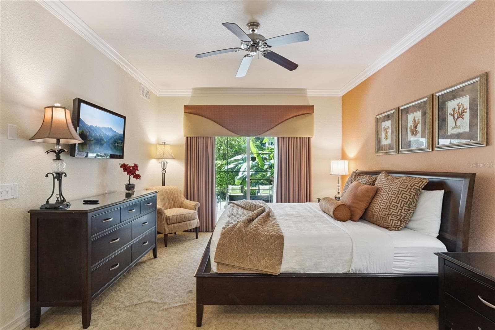 Primary Bedroom: Warmly appointed primary suite featuring crown molding, a ceiling fan, dark wood dresser with wall-mounted TV, cozy seating nook, and sliding glass doors to the outdoor patio. - 7656 WHISPER WAY #102, REUNION