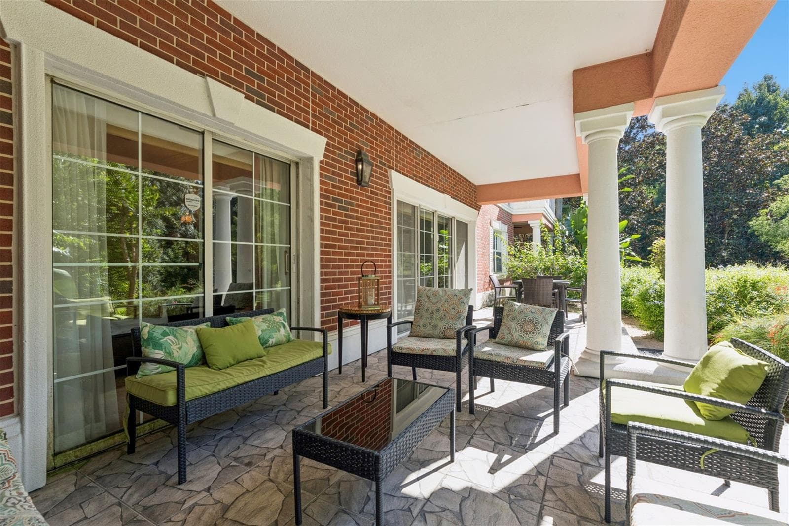 Covered Lanai & Grounds View: Covered porch area featuring white architectural columns, red brick exterior detailing, multiple seating setups, and views of surrounding greenery. - 7656 WHISPER WAY #102, REUNION