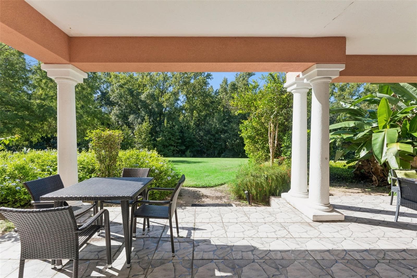 Covered Porch Lawn View: Direct view from the covered patio framed by architectural columns looking out onto an expansive green lawn and dense private tree line. - 7656 WHISPER WAY #102, REUNION