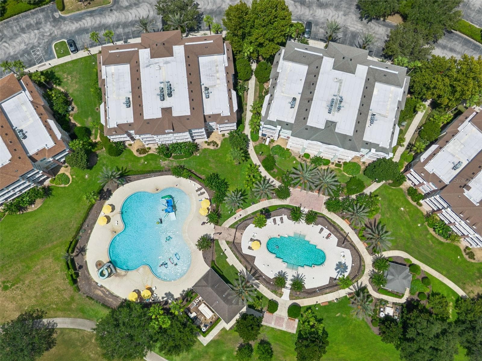 Overhead View of Pool Complex: Direct top-down drone capture showcasing the dual resort pools, splash play area, sunbathing decks, and surrounding condo buildings. - 7656 WHISPER WAY #102, REUNION
