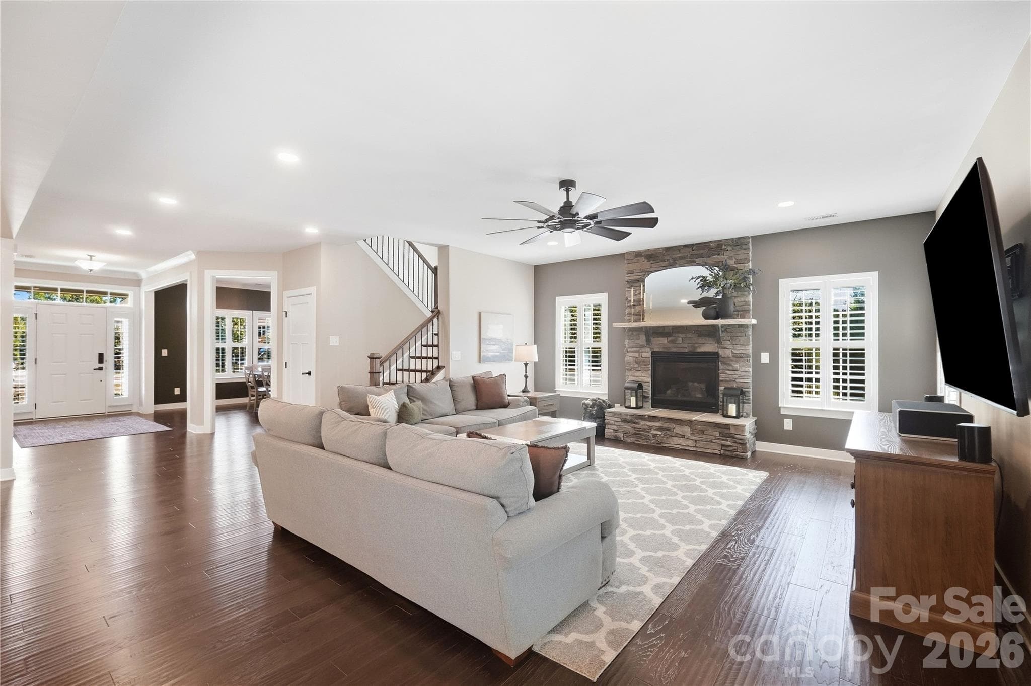 Family room with a stone gas fireplace, plantation shutters, a ceiling fan, and recessed lighting, open to the kitchen, foyer, and staircase. - 912 Manorwood Drive, Waxhaw