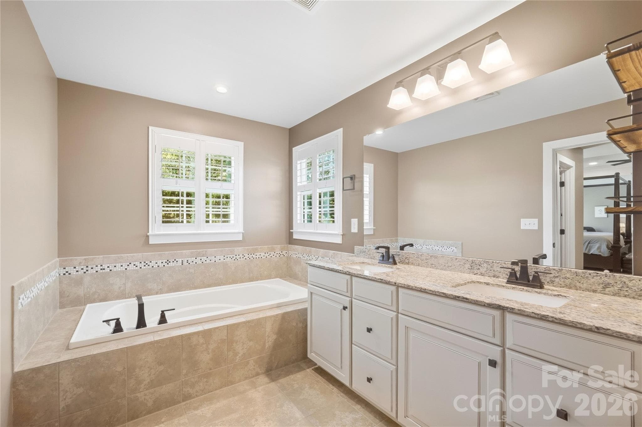 Primary bath with a dual-sink granite vanity, a soaking tub with a tile surround, and plantation shutters. - 912 Manorwood Drive, Waxhaw