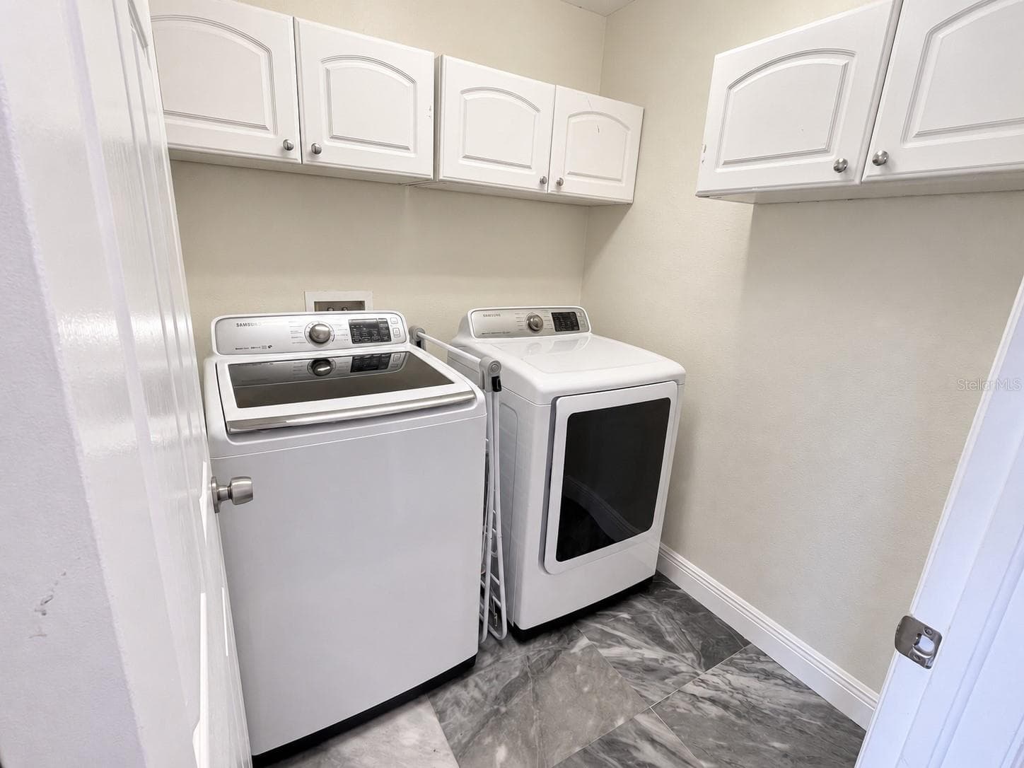 A separate laundry room with side-by-side washer and dryer, overhead storage cabinets, and a tiled floor. - 11117 ANCIENT FUTURES DR, TAMPA