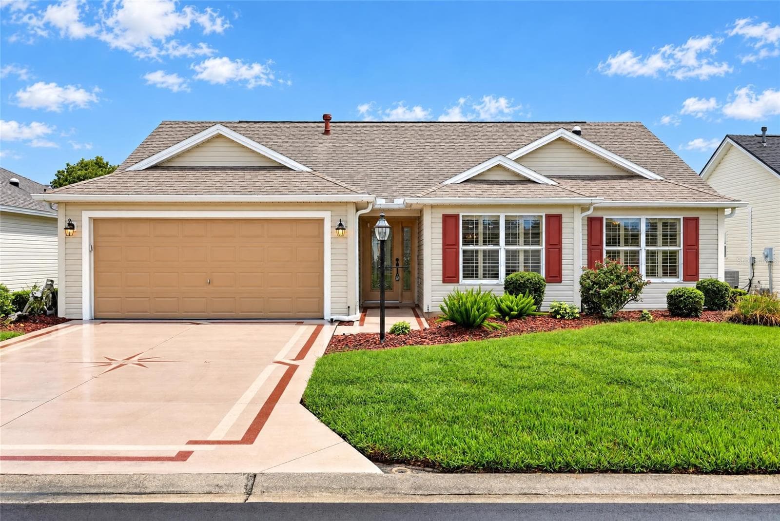 Welcoming front elevation featuring a two-car garage, decorative driveway accents, attractive landscaping, and charming red shutters. - 3064 BURBANK LN, THE VILLAGES