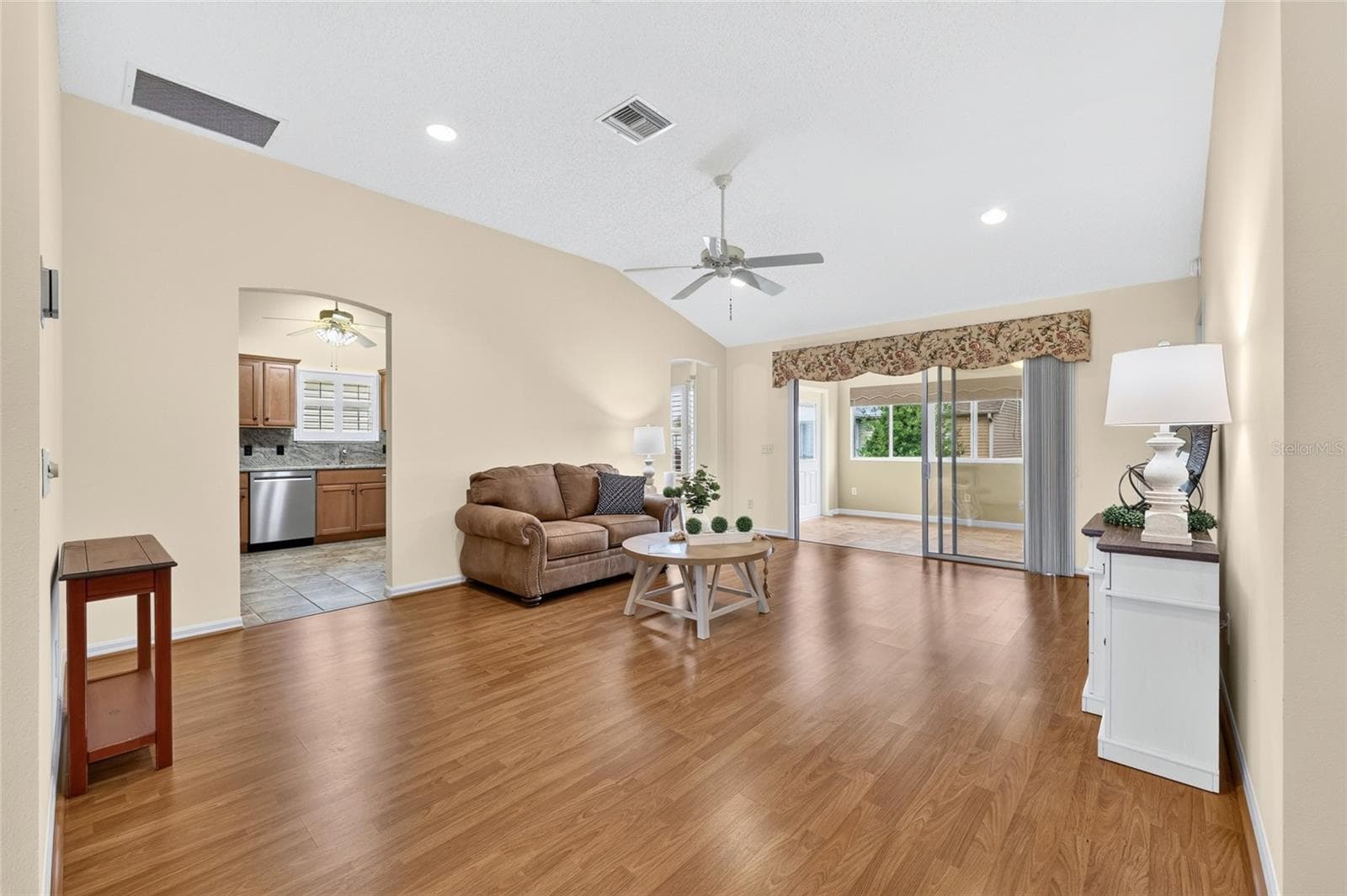 Bright, spacious living room featuring vaulted ceilings, wood-look flooring, recessed lighting, and sliding glass doors leading to the enclosed lanai. - 3064 BURBANK LN, THE VILLAGES