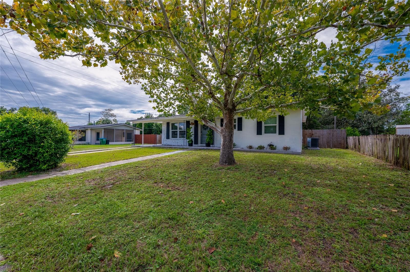 Lush front yard shaded by a large leafy tree, featuring a paved walkway leading to the home's single-story facade with dark window shutters. - 605 E NORMANDY BLVD, DELTONA
