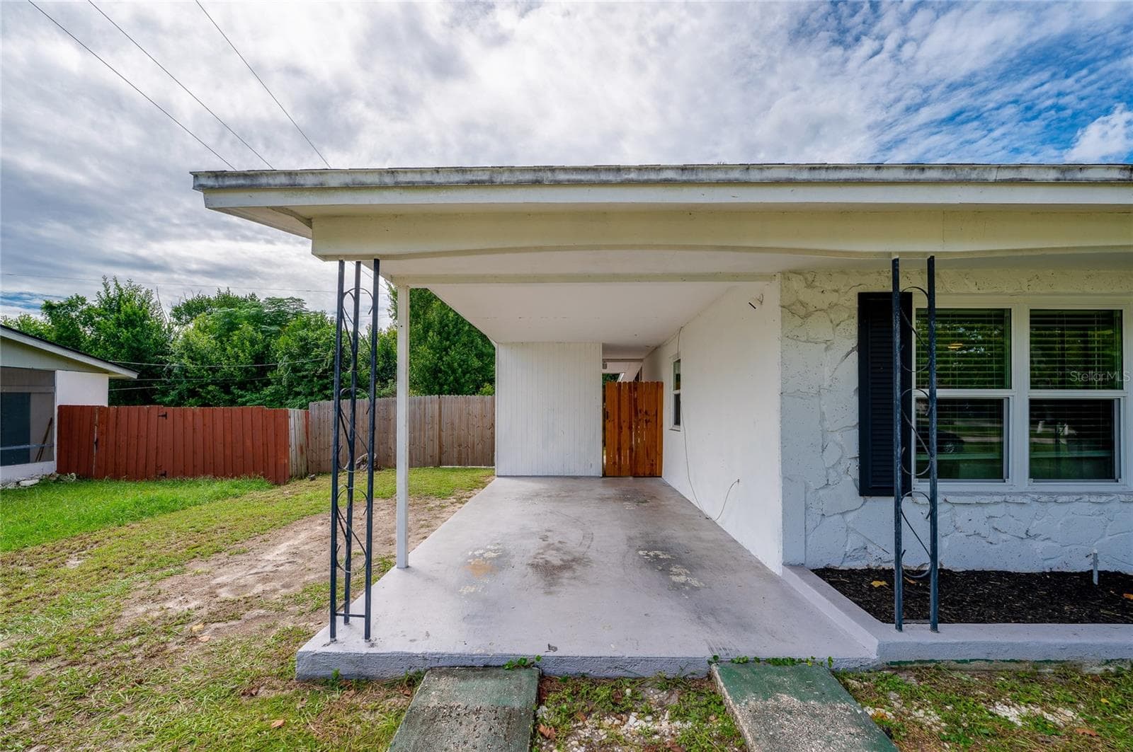 Covered concrete carport with wrought-iron support posts, a textured exterior wall finish, and a gated side pathway. - 605 E NORMANDY BLVD, DELTONA