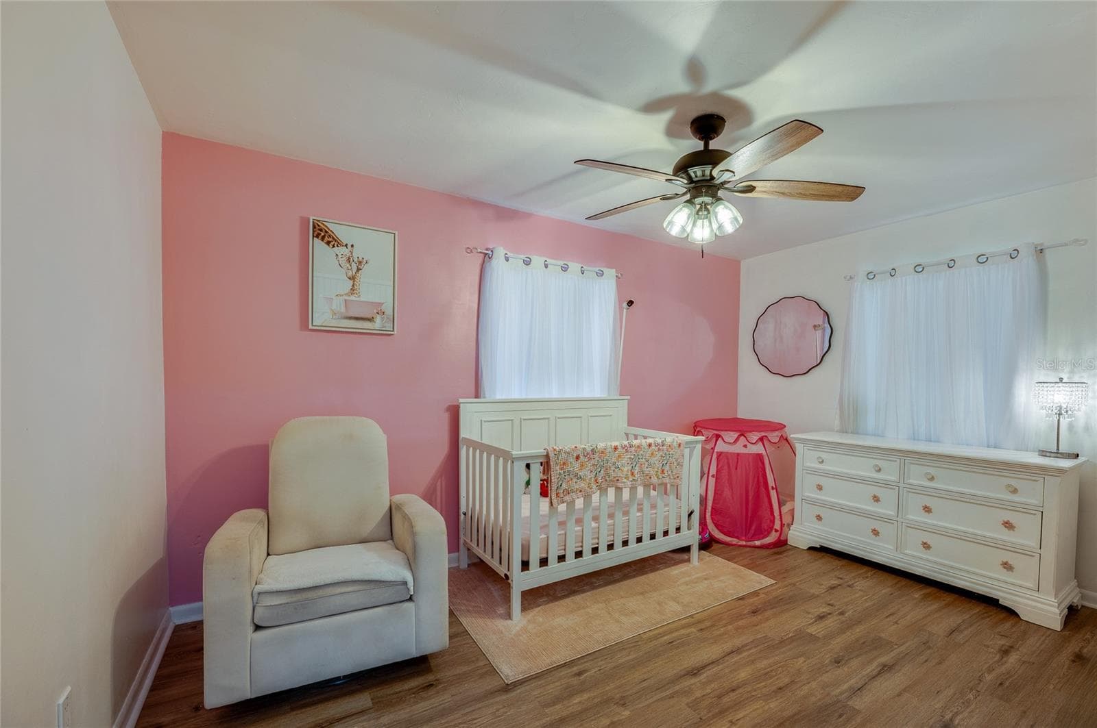 Bright bedroom area highlighted by warm LVP flooring, soft pink accent walls, a ceiling fan, and ample natural light from two windows. - 605 E NORMANDY BLVD, DELTONA