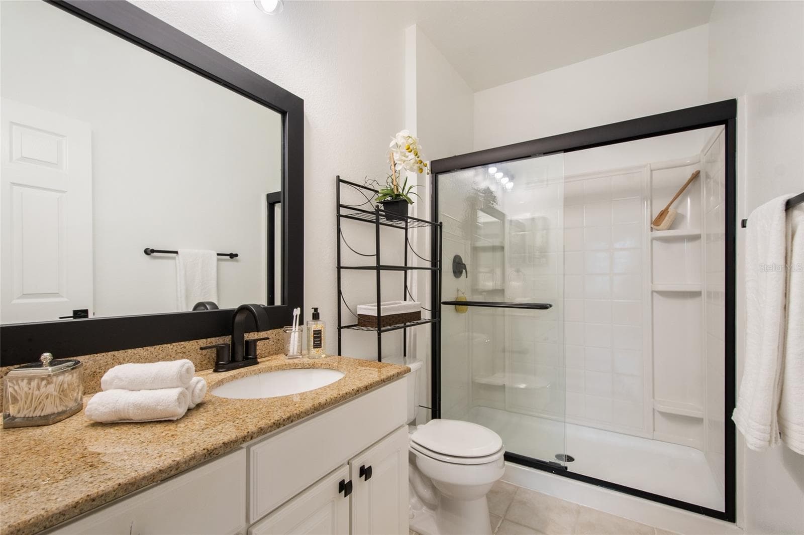 The primary bathroom is totally updated Granite countertops, new moen faucet and shower fixtures, newly framed and painted mirror, new tollet paper holder and completed by a towel bar. The glass shower door is trimmed in black finish to match the balance of the bathroom. Note the new cabinet hardware and the over the toilet storage system that comes with the unit. - 1300 EASTERN PECAN PL #208, WINTER GARDEN