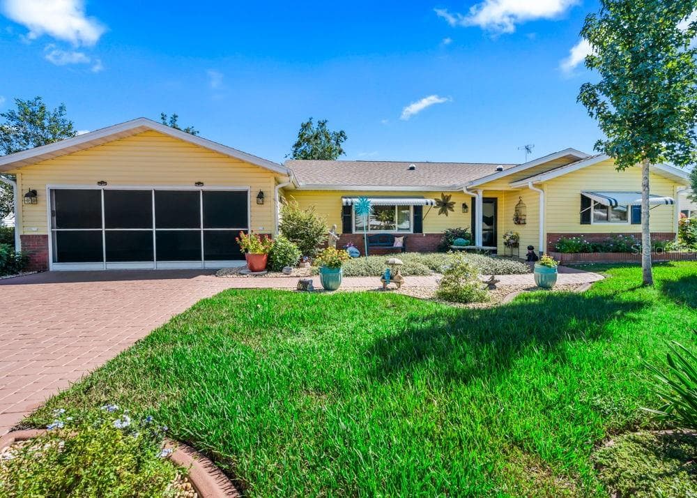 Charming front exterior with freshly painted, cheerful yellow siding, brick accents, a screened garage, and a brick-paver driveway. A covered front porch, striped window awnings, and beautifully landscaped beds filled with colorful flowers that attract butterflies and other pollinators add inviting curb appeal. - 17909 SE 99TH AVE, SUMMERFIELD