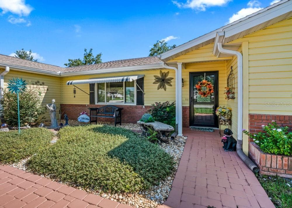 Welcoming covered entry with a brick-paver walkway, cheerful yellow siding, and classic brick accents. Landscaped garden beds, a cozy bench beneath a striped window awning, and decorative touches create a charming first impression. - 17909 SE 99TH AVE, SUMMERFIELD
