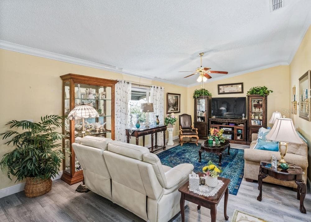 Bright and inviting living room featuring wood-look vinyl flooring, crown molding and a ceiling fan. A large window welcomes natural light, while freshly painted soft neutral walls create a comfortable setting for relaxing or entertaining. - 17909 SE 99TH AVE, SUMMERFIELD